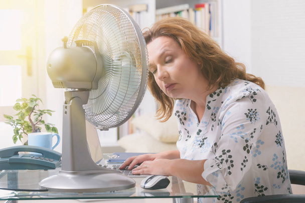 Femme souffre de chaleur tout en travaillant dans le bureau et essaie de se rafraîchir par le ventilateur - Photo, image