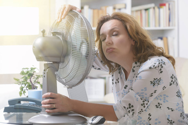 Женщина страдает от жары во время работы в офисе и пытается остыть за счет вентилятора
 - Фото, изображение