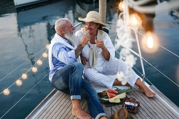 Starsza para toasting szampana i jedzenia owoców na wakacje żaglówka - Szczęśliwi ludzie w podeszłym wieku dobrze się bawią świętując rocznicę ślubu na wycieczce łodzią - Love relationship and travel lifestyle concept - Zdjęcie, obraz