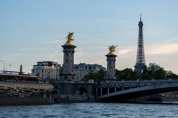 View from seine river to the most ornate, extravagant arch bridge of city Pont Alexandre III after sunset. Towers with gilded Pegasus, fancy Restaurant at riverside and the Eiffel tower in background. - Photo, Image