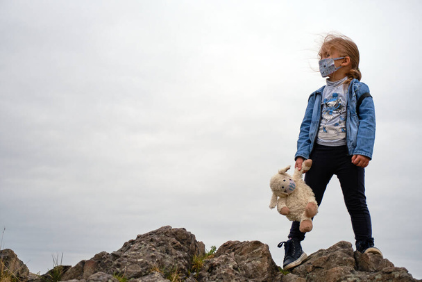 petite fille dans un masque grimpe sur les montagnes avec son jouet dans un masque - Photo, image