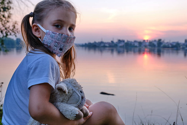 petite fille dans un masque se tient près d'un étang avec son jouet dans un masque derrière le coucher du soleil - Photo, image