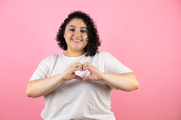 Молодая красивая женщина в повседневной футболке стоит на изолированном розовом фоне улыбаясь в любви показывая символ сердца и форму с руками - Фото, изображение
