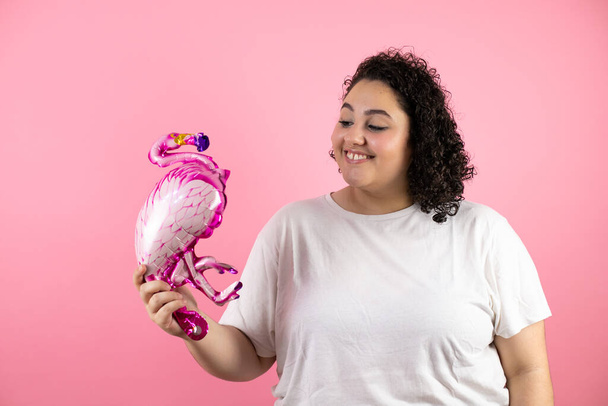 Jovem mulher bonita de pé sobre fundo rosa isolado sorrindo. Olhando e segurando um balão flamengo
 - Foto, Imagem