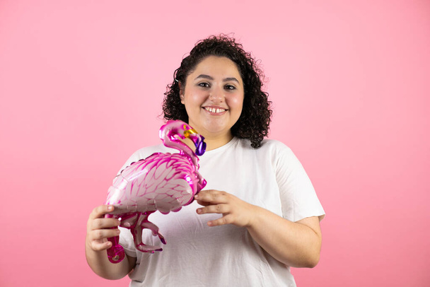 Joven hermosa mujer de pie sobre fondo rosa aislado sonriendo y sosteniendo un globo flamenco - Foto, imagen