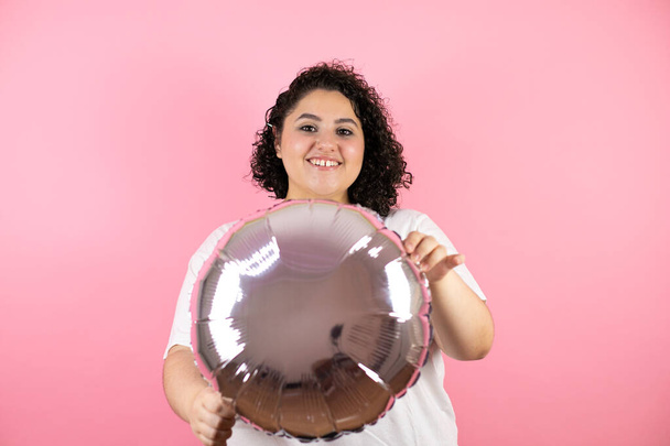 Joven hermosa mujer de pie sobre el fondo rosa aislado sonriendo y sosteniendo un globo - Foto, imagen