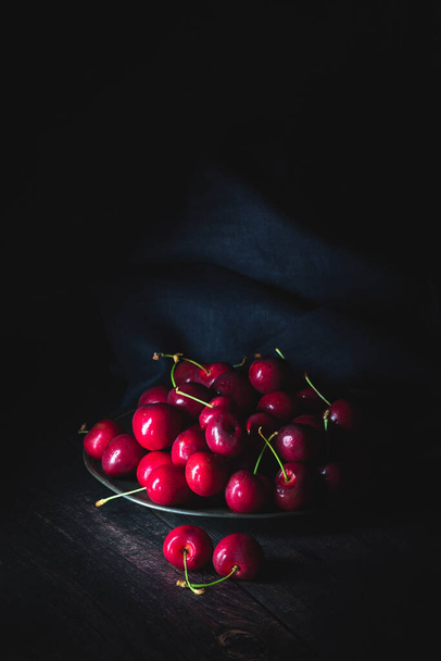 Tummanpunaisia kirsikoita tinalautasella. Heikko valo, pystyssä koostumus. kopiokoneella - Valokuva, kuva