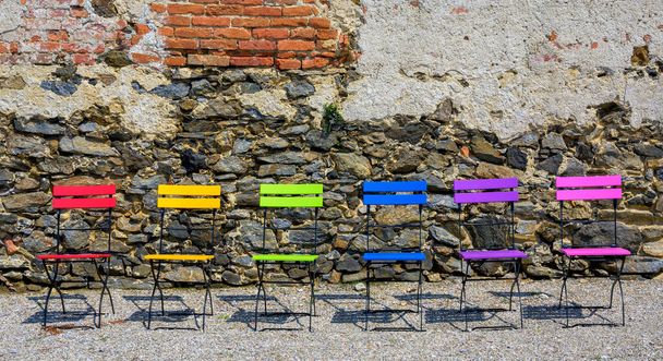 Różne kolorowe składane krzesła przed ścianą z cegły i kamienia z kruszącym się tynkiem w słońcu, Austria - Zdjęcie, obraz
