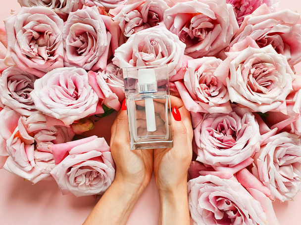 Fehér női kéz piros körmökkel tartsa üveg parfüm és rózsák rózsaszín háttérrel. Eau de wc-t. Mockup, másolás, lapos fektetés. - Fotó, kép