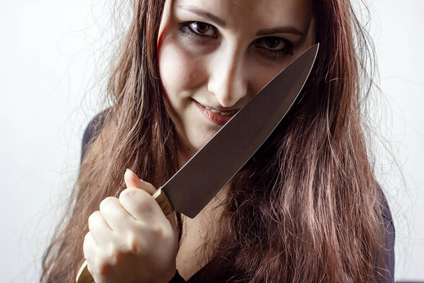 Young caucasian insane brunette woman with knife in black against light background. Halloween concept. - Fotografie, Obrázek