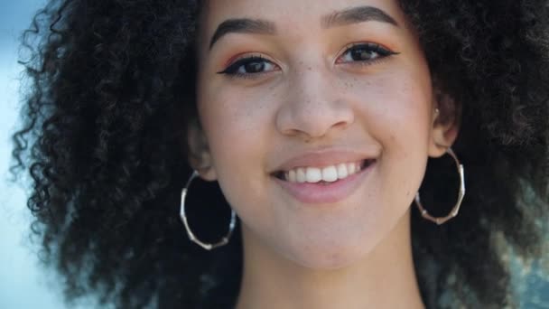 Schöne selbstbewusste junge Afrikanerin lacht. Glückliche ethnische Studentin mit braunen Augen, lockiger Afro-Frisur, runden Ohrringen, leichtem Make-up und Sommersprossen lächelt und zeigt ihre gesunden weißen Zähne - Filmmaterial, Video