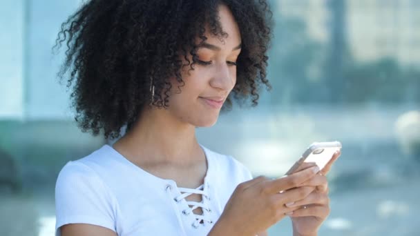 Encantadora chica adolescente afroamericana escribiendo mensaje a un amigo en el teléfono, clics en la pantalla, navegar por Internet. Mujer joven mira a la cámara y sonríe toothy, se ríe de la broma, hace orden en línea en la aplicación. - Metraje, vídeo