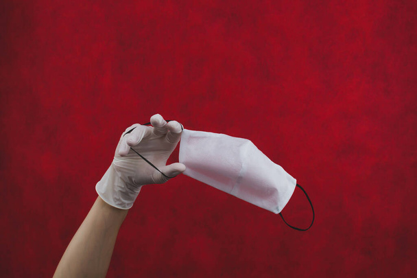 White protective mask hangs on the hand with a white surgical glove. preventive measures against respiratory illness and virus. medical breathing bandage. red background with copy space - Valokuva, kuva