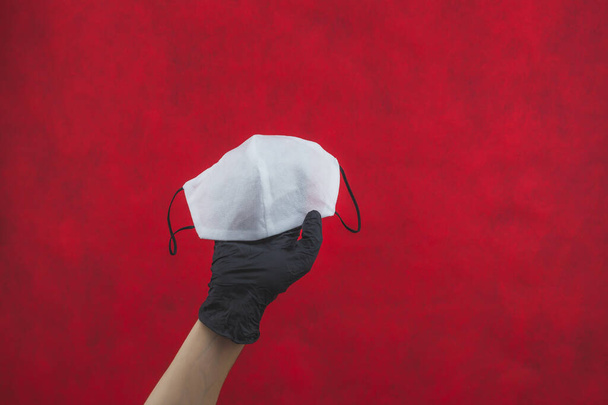 Hand in black surgical glove holds white medical mask on a red background. preventive measures against respiratory illness and virus. disposable masks for protection - Fotoğraf, Görsel