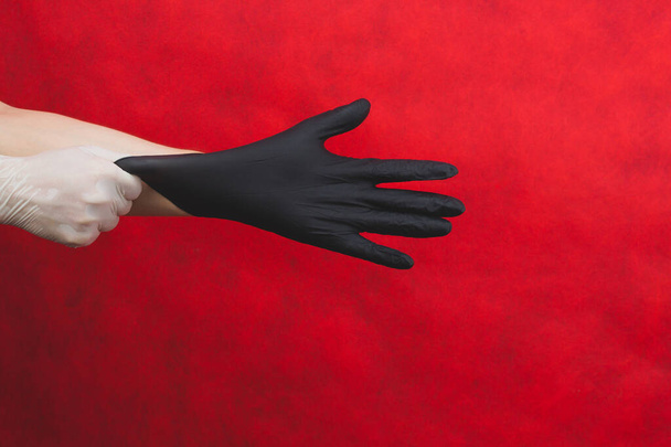 Hand in the white glove puts the black glove on the other. person puts on medical gloves on a red background - Foto, afbeelding