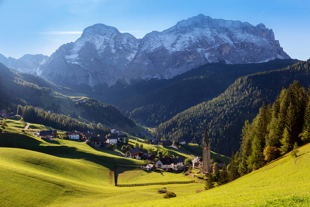 Valle de La Vale - Dolomitas italianas, un pequeño pueblo en las montañas, justo al lado de un bosque alto se encuentra una iglesia esbelta. En el fondo los altos picos de los Dolomitas - Foto, Imagen