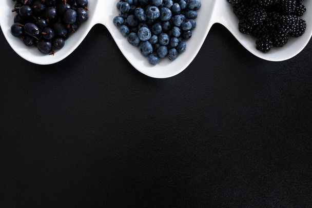 Set of different types of black berries in a white plate on a black table. Stylish seasonal vitamins. - 写真・画像