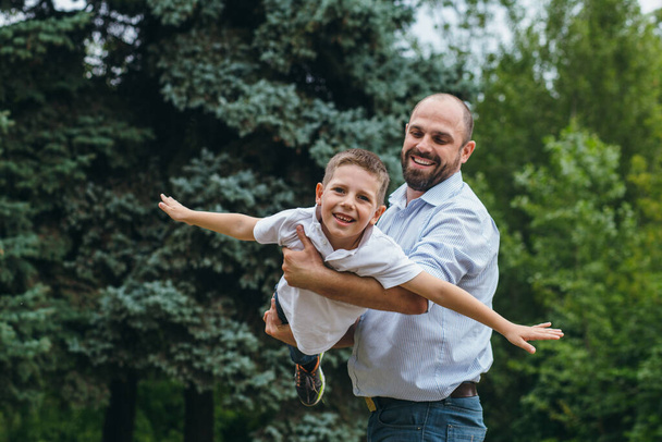Sakallı bir adam parkta genç bir çocuğun omuzlarında yuvarlanıyor. Baba ve oğul doğada uçak oynuyorlar., - Fotoğraf, Görsel