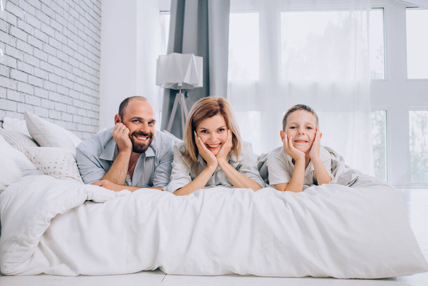 Una familia alegre yace en una cama en un dormitorio blanco y sonríe.  - Foto, imagen