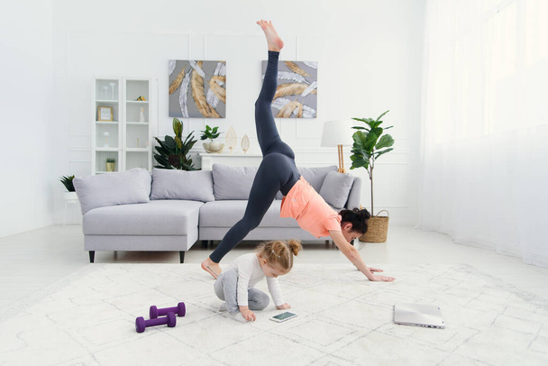 Young adorable mom making stretching exercises and practicing yoga with baby girl at home. Health care and sports concept. - Foto, Bild