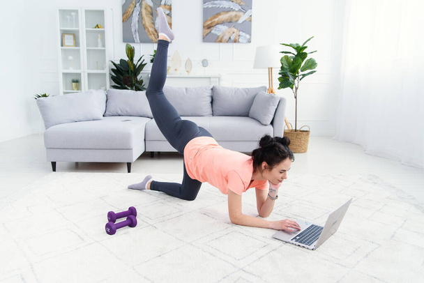 Young sports woman doing workout on the floor at home and watching fitness videos in a laptop. Mom exercising while daughter watching cartoons on background. - Foto, Imagem