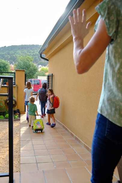 Çocuklar okul için evden ayrılıyor, anne onları garaj yolundan kovuyor, koruyucu maske takıyor. - Fotoğraf, Görsel