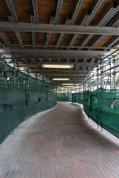 sentier piétonnier à travers un chantier de construction avec des côtés en tissu vert, échafaudage métallique, pavage de briques et éclairage fluorescent - Photo, image