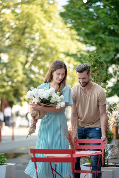 Bearded male pulling red chair for female with bouquet of white flowers, helping her to sit - Фото, зображення