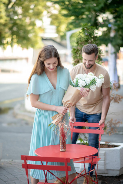 Bearded male pulling chair for female with bouquet of white flowers, helping her to sit down - 写真・画像