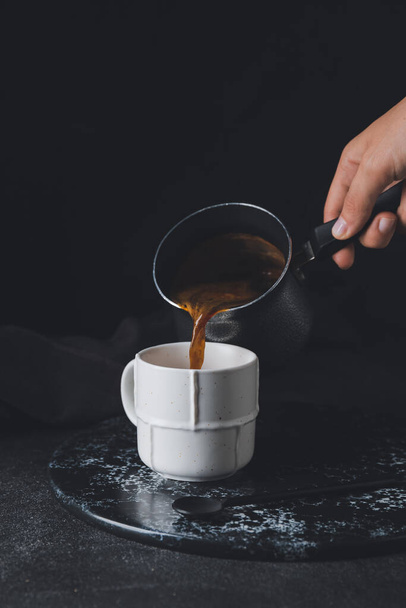 Woman pouring hot coffee from pot into cup on dark background - Fotoğraf, Görsel