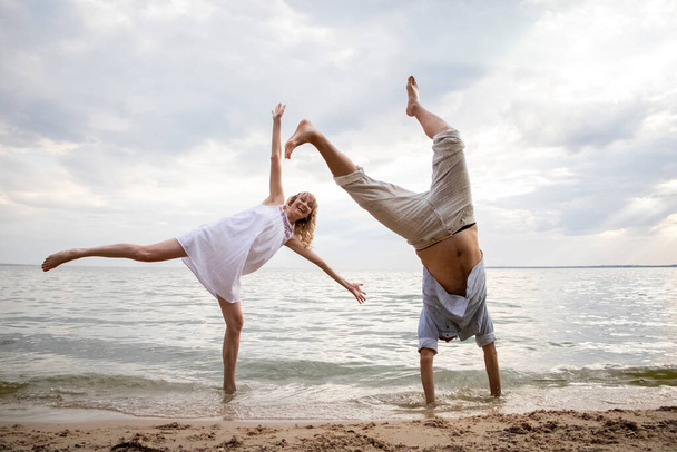 Creative couple relaxing on the ocean beach. A man and a woman do somersaults and jumps in the water. The concept of unusual couples and ingenuity in poses for a photo - Photo, Image
