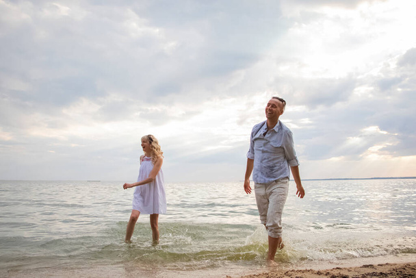Ένας άντρας και μια ερωτευμένη γυναίκα περπατούν στον ωκεανό, γελώντας χαρούμενα και δείχνοντας ευτυχισμένοι. Η έννοια της ευτυχισμένης σχέσης, του ισχυρού γάμου και της οικογενειακής ευτυχίας - Φωτογραφία, εικόνα