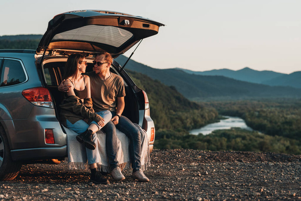 Young Traveler Couple on Road Trip, Man and Woman Sitting on the Opened Trunk of Their Car Over Sunset - Photo, image