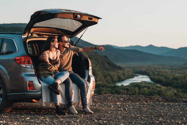Young Traveler Couple on Road Trip, Man and Woman Sitting on the Opened Trunk of Their Car Over Sunset - Photo, Image