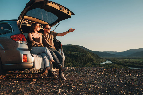 Молодая парочка путешественников в поездке, мужчина и женщина сидят на открытом багажнике своего автомобиля на закате - Фото, изображение