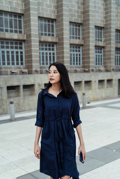 Красивая молодая азиатка в синем платье, выходящая из офиса. - Фото, изображение
