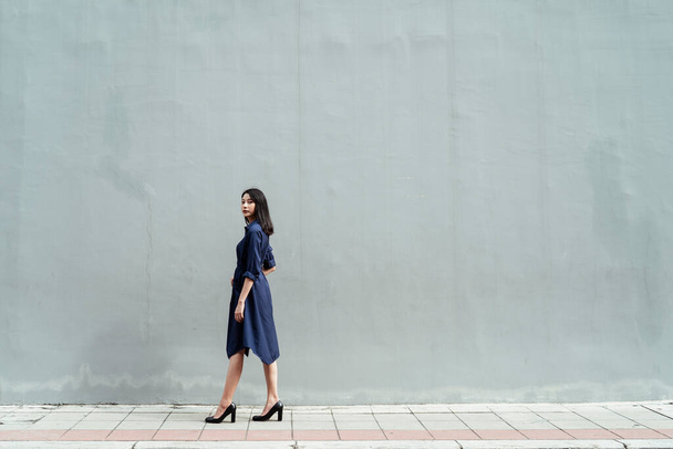 Piękna młoda azjatycka kobieta na niebieskiej sukience stojąca na ulicy przy ogromnej ścianie cementu na tle. - Zdjęcie, obraz