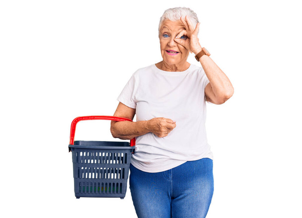 Senior gyönyörű nő kék szemekkel és ősz haj gazdaság szupermarket bevásárló kosár mosolyogva boldog csinál oké jel kéz a szemen keresztül néz az ujjakon  - Fotó, kép