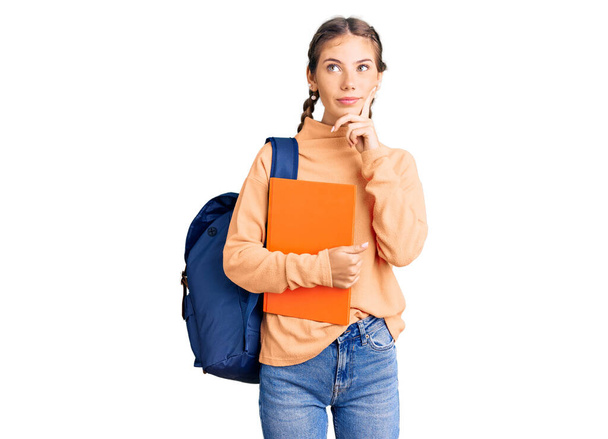 Beautiful caucasian woman with blonde hair wearing student backpack and holding book serious face thinking about question with hand on chin, thoughtful about confusing idea  - Foto, Bild