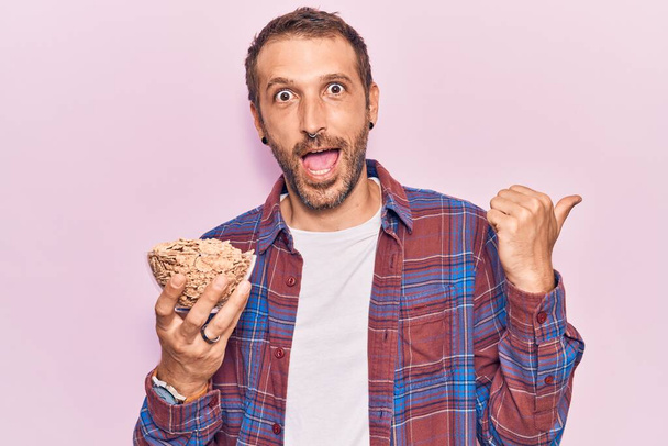 Young handsome man holding cornflakes pointing thumb up to the side smiling happy with open mouth  - Foto, Bild