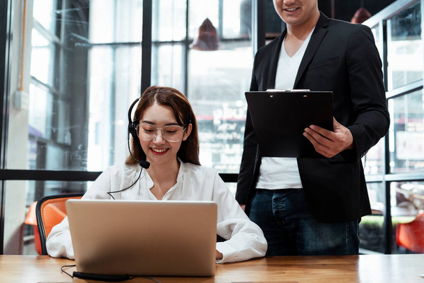 Young asian lady got her job as a call center service, wearing headset with microphone talking with customer, typing on laptop for infomation. Supervisor standing close to her teaching everything about how it work. Trainee. - Foto, Imagen
