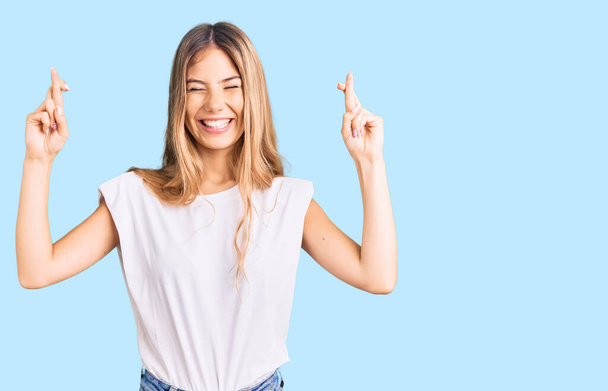 Beautiful caucasian woman with blonde hair wearing casual white tshirt gesturing finger crossed smiling with hope and eyes closed. luck and superstitious concept.  - Photo, Image