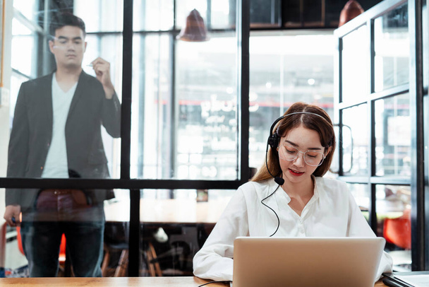 Frau Call Center trägt Headset mit Mikrofon im Gespräch mit Kunden Online-Tippen auf Laptop, während ihr Chef etwas auf Glaswand mit Stift schreibt, Planung. - Foto, Bild