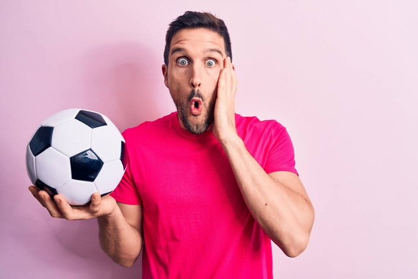 Giovane bel giocatore uomo che gioca a calcio tenendo palla da calcio su isolato sfondo rosa spaventato e stupito con la bocca aperta per sorpresa, faccia incredulo - Foto, immagini