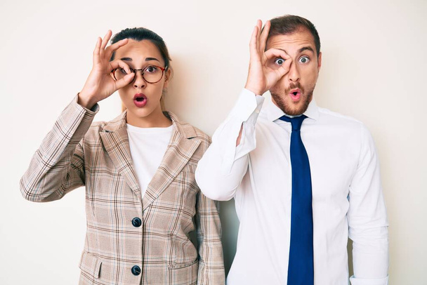 Beautiful couple wearing business clothes doing ok gesture shocked with surprised face, eye looking through fingers. unbelieving expression.  - Photo, Image