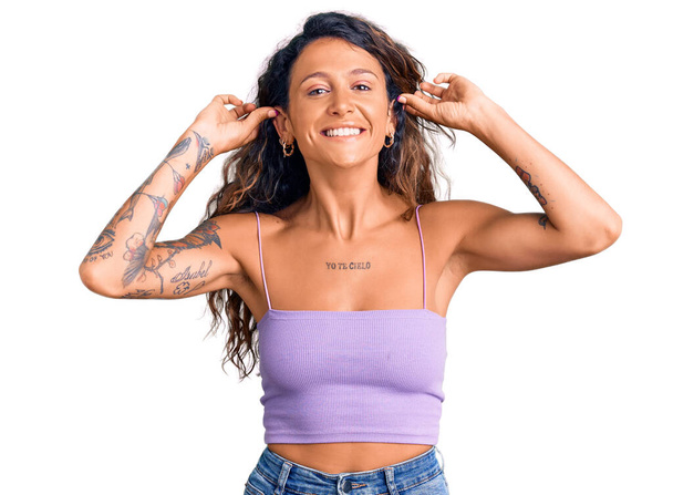 Giovane donna ispanica con tatuaggio che indossa abiti casual sorridente tirando le orecchie con le dita, gesto divertente. problema di audizione  - Foto, immagini