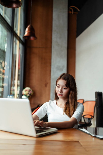 Azji młoda kobieta na białej sukience koncentruje się pracy na laptopie w nowoczesnym pokoju biurowym. - Zdjęcie, obraz