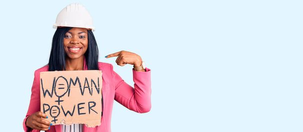 Jovem afro-americana vestindo capacete arquiteto segurando mulher poder banner apontando dedo para um auto sorrindo feliz e orgulhoso
  - Foto, Imagem