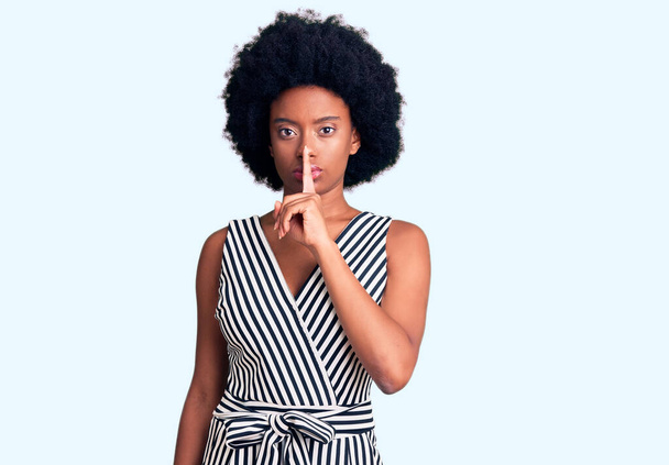 Junge afrikanisch-amerikanische Frau in lässiger Kleidung, die darum bittet, mit dem Finger auf den Lippen ruhig zu sein. Schweigen und geheimes Konzept.  - Foto, Bild