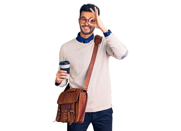 Mladý pohledný hispánec muž v kožené tašce a pití vzít kávu usměvavý šťastný dělá ok znamení s rukou na oku dívá skrz prsty  - Fotografie, Obrázek
